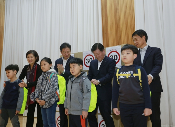 현대위아 김경배 사장(두 번째 줄 오른쪽 두 번째)이 20일 경남 창원시 성산구 안민초등학교에서 열린 ‘현대위아 어린이 교통안전 지킴이 캠페인’에서 어린이용 안전 가방 덮개를 한 학생의 가방에 씌우고 있다. (사진=현대위아)